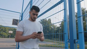 Young Middle eastern Arabic athlete in summer sportswear walking moving along stadium fence holding mobile phone, using online app on smartphone, texting, chatting typing message, touching screen