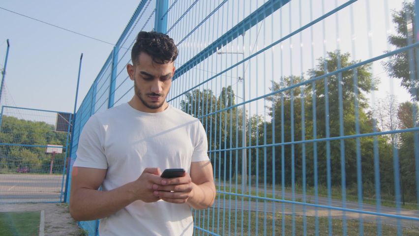 Young Middle eastern Arabic athlete in summer sportswear walking moving along stadium fence holding mobile phone, using online app on smartphone, texting, chatting typing message, touching screen | Shutterstock HD Video #1079962088