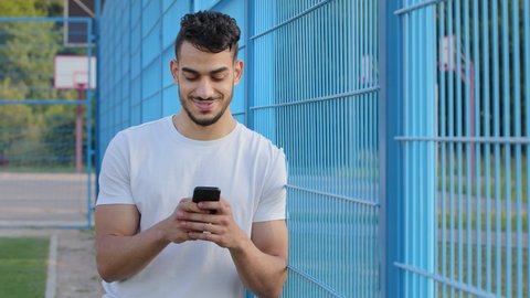 Middle eastern indian man holding phone in hands texting message or using mobile apps, checking social media applications on smartphone. Young Arabic athlete in summer sportswear playing on cellphone