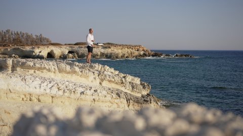Extreme wide shot of happy male tourist throwing rocks in blue Mediterranean sea water in slow motion standing on rocky coast. Side view of Caucasian man with Japanese respect and Power tattoos