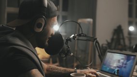 Tracking shot of cheerful man in headphones playing video game and talking into microphone while streaming online from home or studio