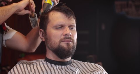 Close-up of a handsome Caucasian guy sitting in a hairdressing salon. An unrecognizable barber parses hair with a comb. 4K 50 fps slow motion