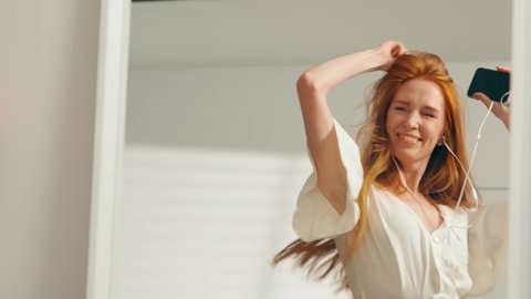 Portrait close-up: A young attractive girl with red hair is dancing merrily at home in front of a mirror with headphones. A beautiful woman in a white dress is dancing with her hands up. Slow mo, 4K