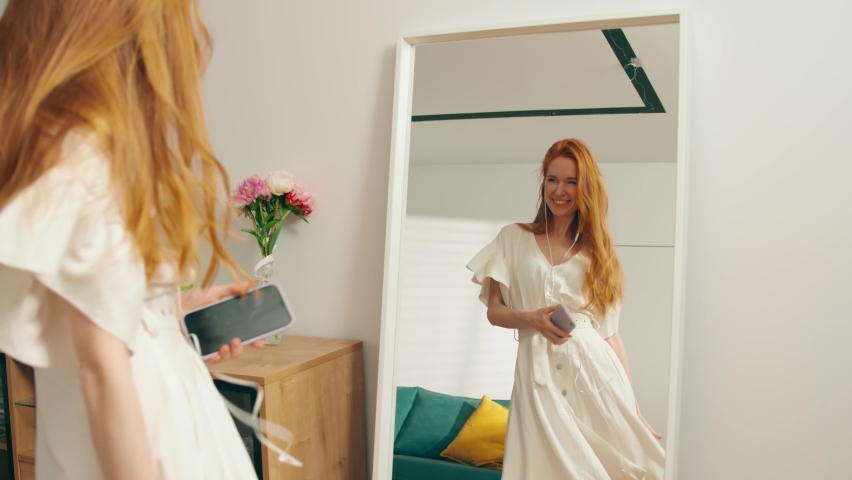 The general plan: A young attractive girl with red hair is dancing merrily at home in front of a mirror with headphones. A beautiful woman in a white dress admires herself touching hair. Slow mo, 4K Royalty-Free Stock Footage #1079975867