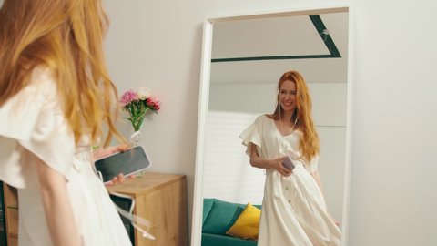 The general plan: A young attractive girl with red hair is dancing merrily at home in front of a mirror with headphones. A beautiful woman in a white dress admires herself touching hair. Slow mo, 4K