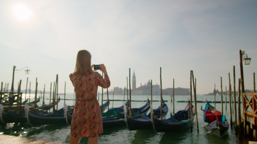 Back view of Lady Taking Photos by Phone with Gondolas in Venice Grand Canal, Italy, Europe. Young Female Tourist or Blogger. Travel Blogging and Landmark Sightseeing Concept. 4K zoom in gimbal shot