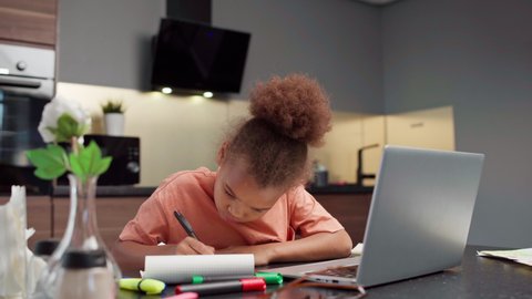 Biracial little girl doing homework at home. Kid writing in notepad and struggling with difficult task. Open laptop pc on table. Distance education and online learning at quarantine concept