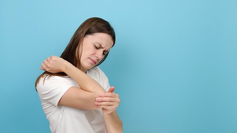 Close up of unhappy sad young caucasian woman 20s old years having pain in his elbow, warming up injured hand after trauma, posing isolated over blue color background wall studio with copy space