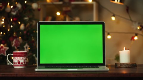 Laptop with green screen close-up. Computer with chroma key standing on table, burning candle and red cup. Christmas tree on background. New Year holidays concept. 