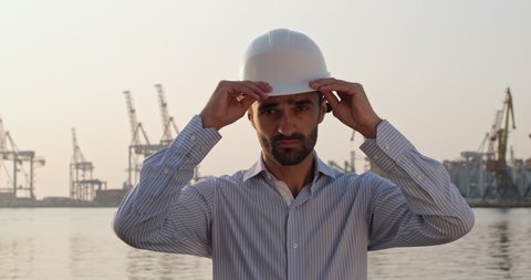 A man in a shirt puts a protective white helmet on his head while standing in the seaport against the background of cargo cranes. Man in charge of affairs at the seaport.