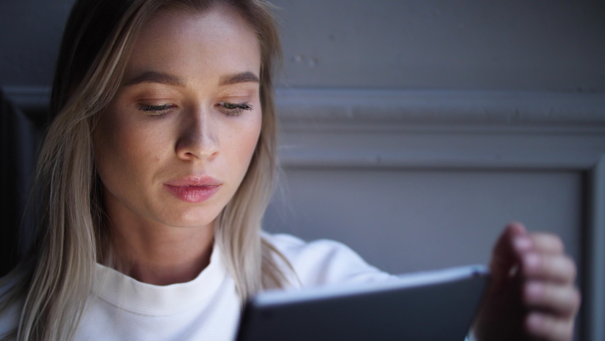 Portrait of lonely blonde woman sitting in cozy room with ipad tablet computer device. Female touching, tapping on screen surface. Browsing, surfing internet applications via portable table computer.