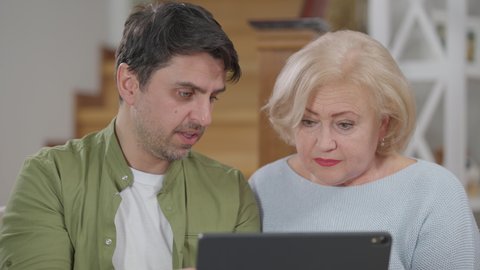 Portrait of confused senior woman and patient adult man talking pointing at tablet. Calm confident handsome Caucasian son teaching embarrassed mother using modern technologies in daily routine