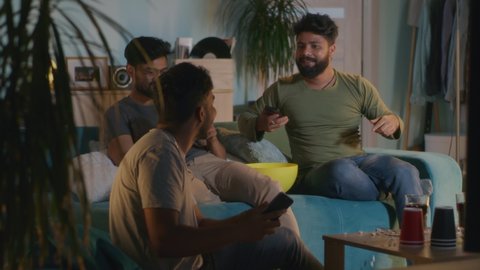 Indian men with cellphones talking about online stakes and watching sports competition together while gathering in living room in evening at home