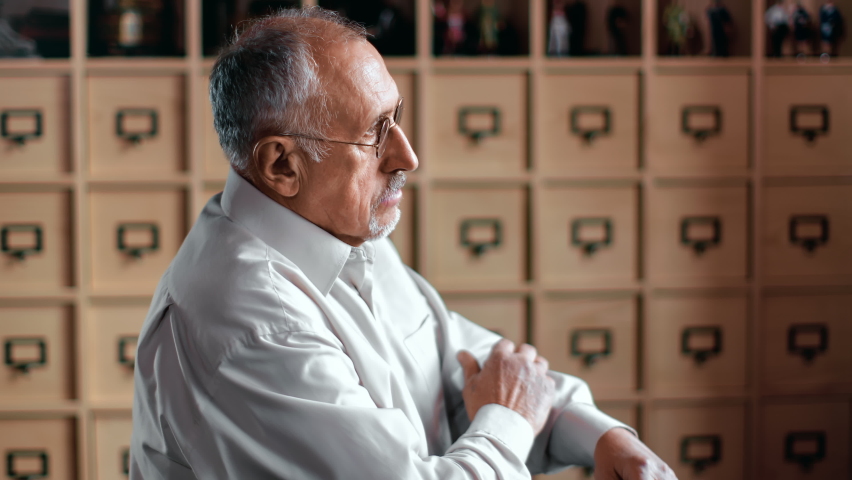 Closeup pensive elderly man standing with crossed hands at library archive bookshelves storage side view. Focused 70s professor scientist writer author thinking about historical antique researching
