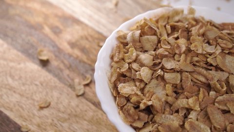 Dry uncooked spelt flakes fall into a white bowl on a wooden rustic background in slow motion. Close up. Healthy natural food concept