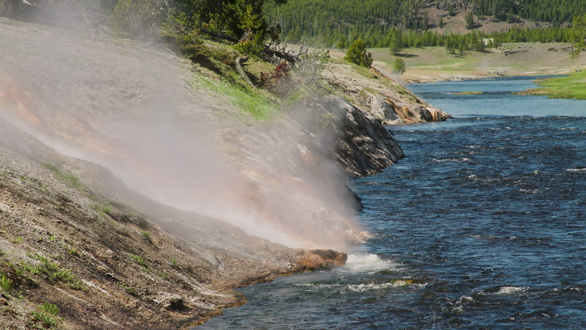 Volcanic nature wilderness on sunny summer day with green forest background. World famous Yellowstone National Park 4K cinematic landscape with steaming hot spring stream flowing into deep blue river