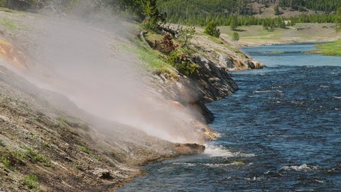 Volcanic nature wilderness on sunny summer day with green forest background. World famous Yellowstone National Park 4K cinematic landscape with steaming hot spring stream flowing into deep blue river
