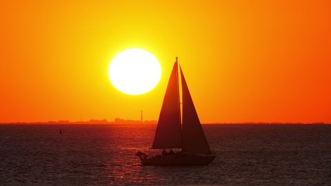 Small yacht with all sails set pass by at gulf, silhouetted shot against sunset, big sun disc hanging low over horizon line. Scenic telephoto shot of sunset at sea shore