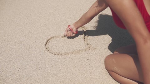 Shape of heart drawn on the sand. Girl sitting on the sand drawing symbol of love. Silhouette of young woman in red swimsuit in nature, depicting heart of sand. The concept of recreation and nature