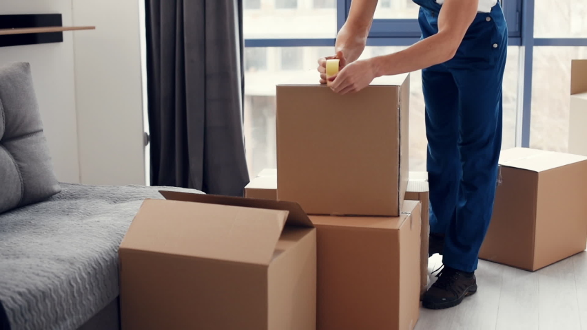 Packaging the box. Young male mover in blue uniform works indoors in the room. Royalty-Free Stock Footage #1080016208