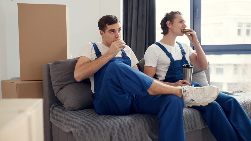 Tired and taking break. Two young movers in blue uniform working indoors in the room.