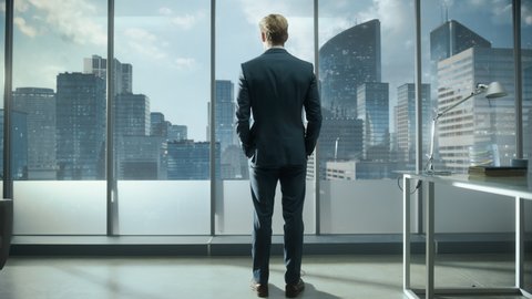 Successful Young Businessman in a Perfect Tailored Suit Standing in His Modern Office Looking out of the Window on Big City with Skyscrapers. Successful Finance Manager Planning Project Strategy.