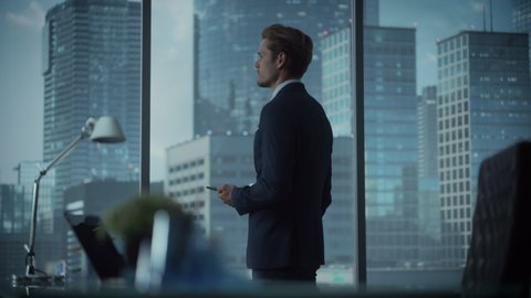 Successful Young Businessman in a Suit Standing in His Modern Office, Using Smartphone, Looking out of the Window on Big City with Skyscrapers. Successful Finance Manager Planning Work Projects.