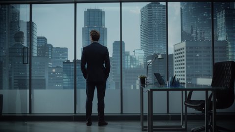 Confident Young Businessman in a Perfect Tailored Suit Standing in His Modern Office Looking out of the Window on Big City with Skyscrapers. Successful Finance Manager Planning Project Strategy.