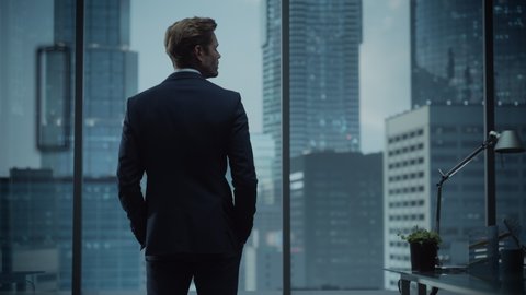 Thoughtful Young Businessman in a Perfect Tailored Suit Standing in His Modern Office Looking out of the Window on Big City with Skyscrapers. Successful Finance Manager Planning Project Strategy.
