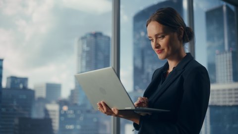Successful Businesswoman in Stylish Dress Working on Laptop, Standing Next to Window in Big City. Confident Female CEO Analyze Financial Projects. Manager at Work Planning Marketing Campaign.