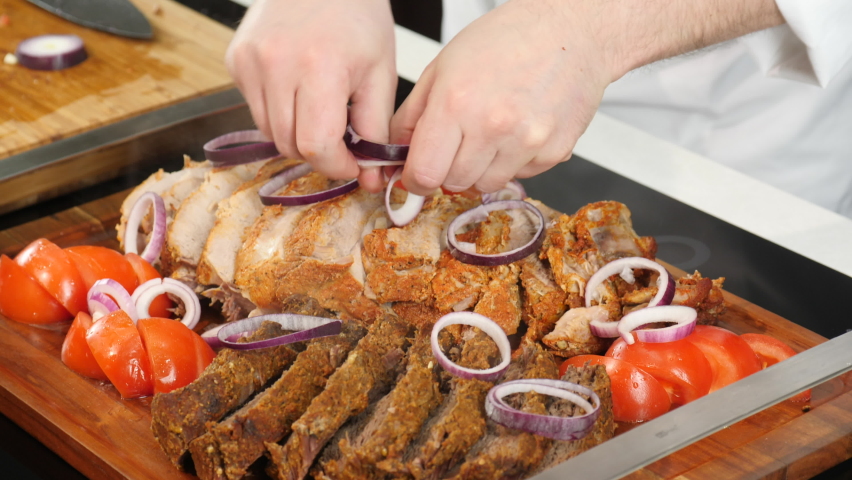 Chef cook in restaurant seasons roasted or grilled pork, beef chop slices with onion rings and tomatoes. BBQ tenderloins, kebab sirloins on wooden tray. Meat steaks vegetable ingredients flavoring.
