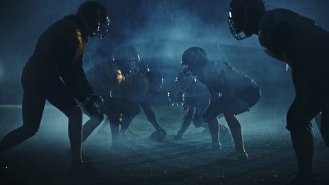 American Football Field Two Teams Compete: Players Pass and Run Attacking to Score Touchdown Points. Cinematic Rainy Night with Athlete Silhouettes Fight for the Ball in Dramatic Fogey Slow Motion