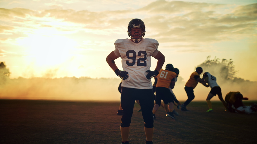 American Football Game: Portrait of a Tough Muscular Player Wearing Helmet, Posing, Looking at Camera. In Background Teams of Professional Athletes Compete for Championship Victory. Cinematic Handheld