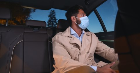 Middle-eastern man in protective face mask sitting on backseat in taxi. Portrait of young businessman wearing safety mask riding on backseat in car late at night
