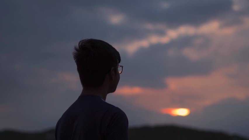 Silhouette of an Indian kid looking at the orange sky during the sunset. Hopeful kid stares at the sun. Kid lost in deep thoughts after looking at the sunset.  | Shutterstock HD Video #1080025601