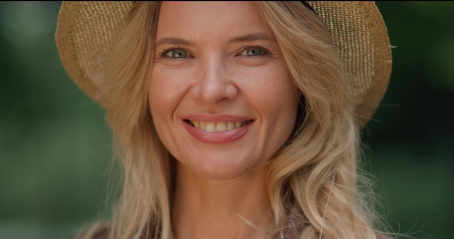 Portrait of Smiling Beautiful Adult Caucasian Blond Woman in Straw Hat. Pretty Middle-Aged Lady with Blue Eyes and Attractive Appearance looking into camera. Standing outdoors. Sincere Emotions. | Shutterstock HD Video #1080026249