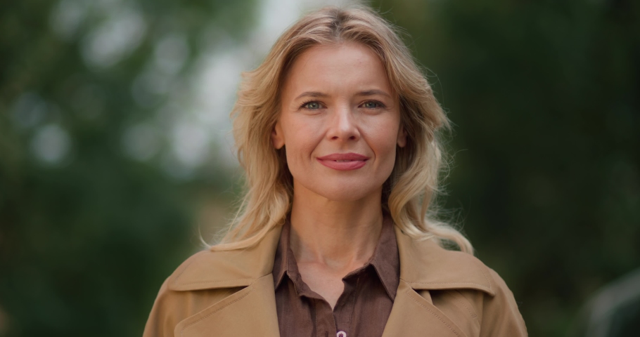 Portrait of Amazing Caucasian Blond Middle-Aged Woman with Beautiful Blue Eyes. Smiling Adult Elegant Lady wears Coat standing outdoors while looking into camera. Pretty Appearance of Mature Female.