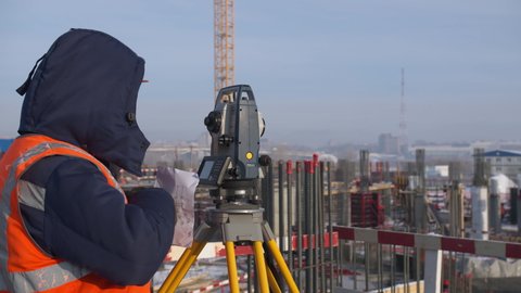 OMSK, RUSSIA - FEBRUARY 04 2021: Engineer in bright vest and warm jacket uses theodolite to measure ground angle at construction site on winter day on February 04 in Omsk