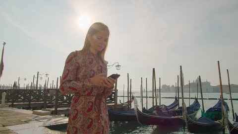 Millennial Woman Uses SmartPhone App Walking in Venice Grand Canal with Gondolas at Summer Sunrise. Mobile Internet Web Surfing or Texting Concept. 4K gimbal follow medium wide shot