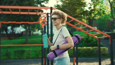 Mature woman going to yoga class outdoors. Active senior enjoying healthy lifestyle. Senior female holding exercise mat while walking at training session. Positive elderly woman with sports equipment