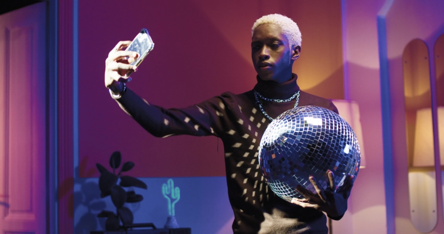Young stylish African American man taking selfie with disco ball for social networks using camera smartphone while standing on retro party.