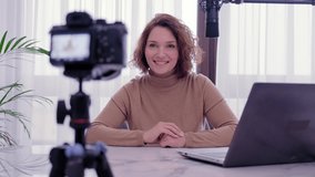 Woman blogger videotapes her vlog at home. Smiling woman recording her video blog. Adult woman speaks in front of a video camera for her blog channel. Vlogger makes online streaming using smartphone