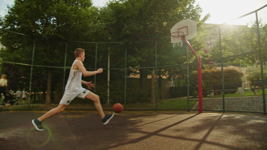 Footage of a teenager dribbling on an outdoor court, during a bright and sunny summer day. Video of a white caucasian teen training to play basketball. Video of a male doing basketball tricks. 