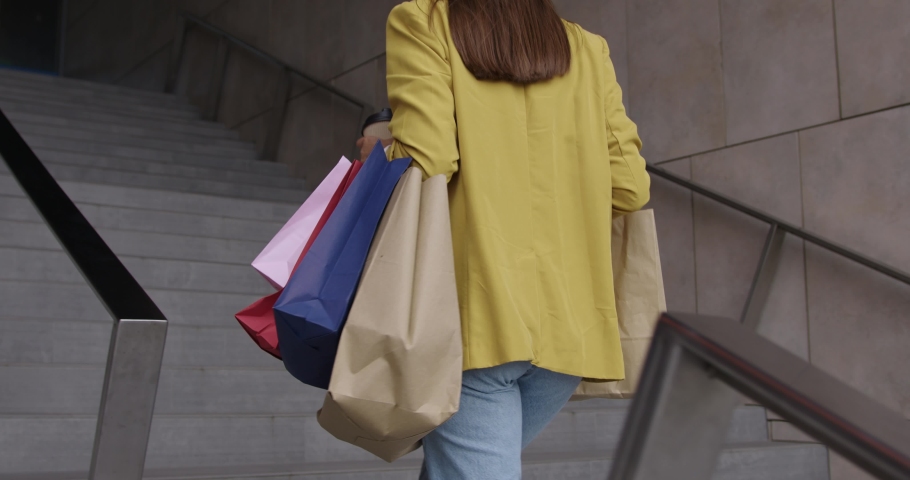 Close up of young woman in blue jeans and yellow jacket walking up on stairs with colorful shopping bags in hands. Concept of people, sales and black friday.