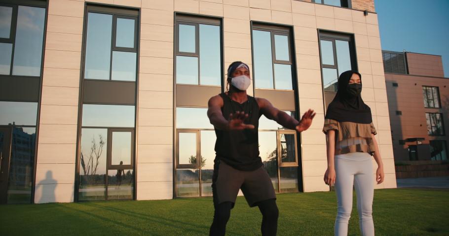 Muslim family of black man and Asian woman with protective masks does squats at training near building with mirror windows slow motion