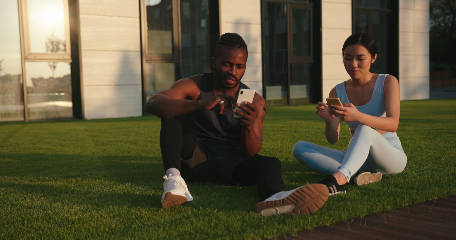 Asian woman and African-American man friends use mobile phones resting after training on lawn grass at sunset slow motion