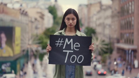 Young woman with brown hair standing on city street and holding banner with hashtag Me Too. Asian female supporting movement against harassment and sexual assault.