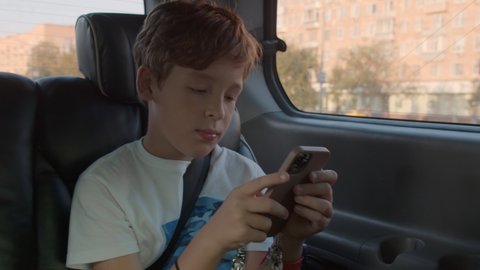 Teen boy surfing net or social media on the phone while traveling by car and being bored with the ride. Child is fastened with seat belt