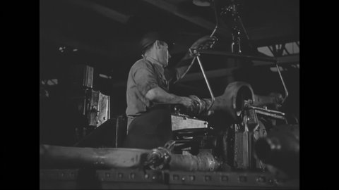1930s: Worker lifts rear axle from drill press and places on rollers, then moves another piece into the drill. Worker uses tool to finish the axle. Slate. Piece placed in drill.