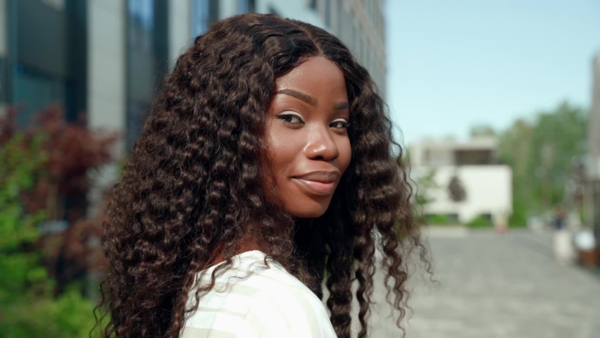 Happy smiling young ethnic black African attractive millennial business woman or student with curly long hair standing outdoor on sunny city street looking at camera. Slow motion close up portrait.